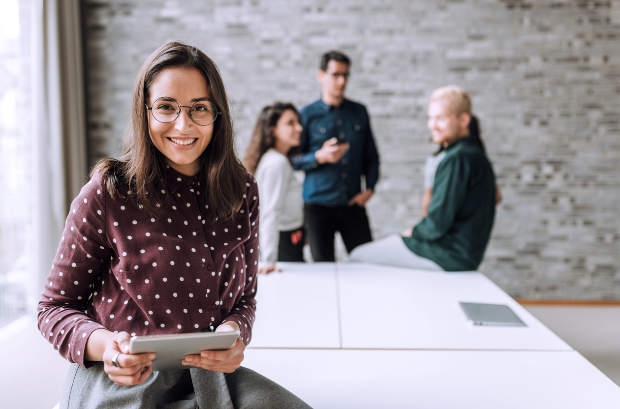 Mujer emprendedora sonriente delante de una mesa de reuniones con grupo de jóvenes emprendedores al fondo.