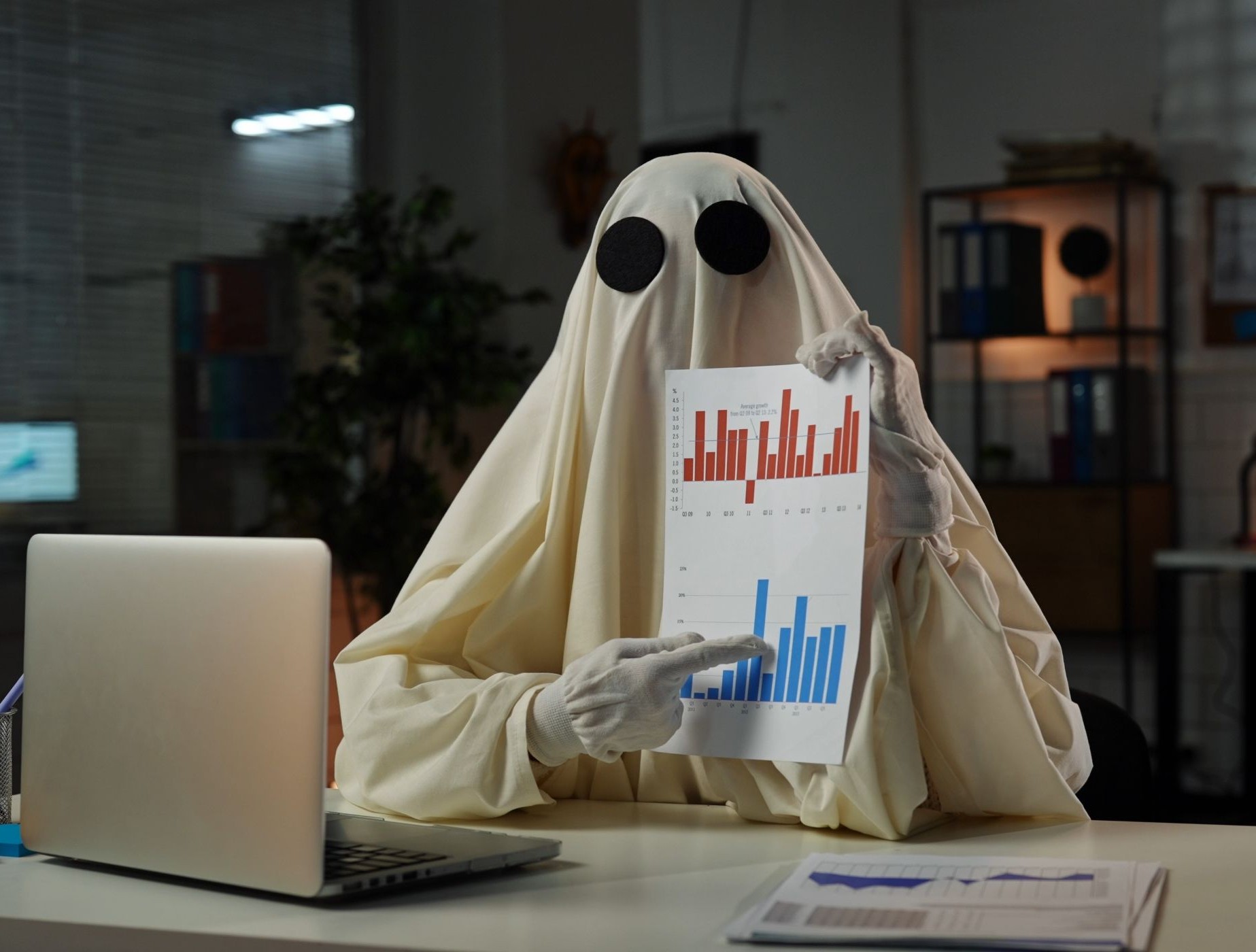Empresario con sábana como fantasma mostrando gráficos ante un escritorio sobre el que hay un ordenador portátil